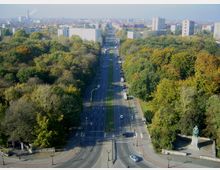 Blick auf eine breite Straße, die von Bäumen gesäumt und durch einen mittleren Grünstreifen getrennt wird, mit umliegenden Wohngebäuden und Parks im Hintergrund. Im Vordergrund ist eine Kreuzung mit Gehwegen und eine Statue auf einem kleinen Platz zu sehen.
