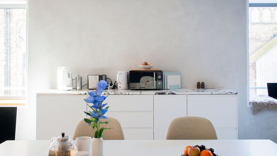 Heller Küchen- und Essbereich mit weißen Unterschränken und marmorierter Arbeitsplatte, auf der kleine Küchengeräte wie Wasserkocher, Toaster und Mikrowelle stehen. Im Vordergrund ein weißer Esstisch mit Teekanne, Vase mit blauen Blumen und einer Obstschale; links und rechts sorgen große Fenster für Tageslicht.