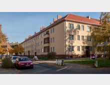 Mehrstöckiges Wohngebäude im Stil der 1930er Jahre mit gelber Putzfassade, rotem Ziegeldach und dunkelroten Klinkerdetails. Die Straße im Vordergrund ist von parkenden Autos und herbstlich gefärbten Bäumen gesäumt.