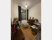 Ein Wohnzimmer oder Arbeitszimmer mit einem großen Fenster, das von dunklen Vorhängen eingerahmt ist. Im Raum befinden sich ein Sessel, ein Schreibtisch mit Büromaterialien, eine Holzkommode, ein Kleiderständer und weitere Möbel auf einem Parkettboden.