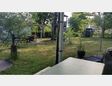 Garten mit Rasenfläche, Bäumen und Sträuchern, im Vordergrund eine alte Handwasserpumpe und ein gedeckter Tisch. Im Hintergrund sind eine Pergola mit Sitzgelegenheiten, ein Spielturm aus Holz und ein geparktes Fahrzeug zu sehen.