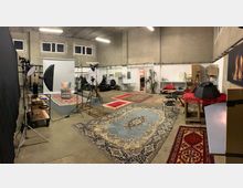 Großes Fotostudio mit hohen Betonwänden und mehreren fotografischen Ausrüstungsgegenständen wie Softboxen, Stativen und Kulissen. Der Boden ist mit verschiedenen orientalen Teppichen bedeckt, im Hintergrund sind weiße Wände mit Regalen, Möbeln, einer Pflanze und einem kleinen Tisch sichtbar.