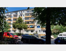 Mehrstöckiges Wohngebäude mit Balkonen, teils mit Sonnenschirmen ausgestattet, umgeben von Bäumen und geparkten Autos. Im Vordergrund befinden sich ein roter Lieferwagen, ein parkender Kombi sowie Fahrräder.