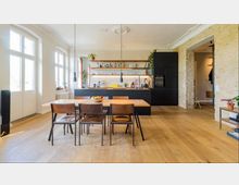 Moderne Wohnküche mit einem großen Holztisch und sechs Stühlen im Vordergrund, einer schwarzen Küchenzeile mit offenen Regalen und Hängelampen im Hintergrund. Die Wände aus weißem Putz und teilweise sichtbaren Ziegeln sowie der helle Holzboden prägen die Gestaltung.