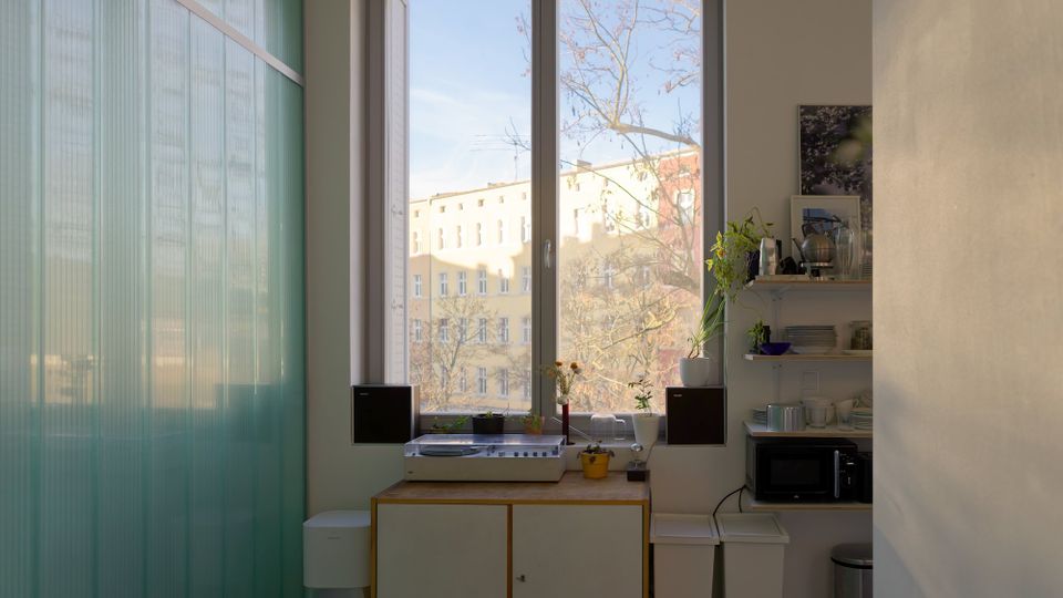 Heller Innenraum mit großem doppelflügeligem Fenster und Blick auf Wohngebäude und kahle Bäume. Unter dem Fenster stehen ein Sideboard mit Plattenspieler und Pflanzen sowie rechts offene Regale mit Geschirr und einer Mikrowelle; links eine halbtransparente grünliche Glaswand.