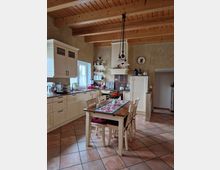 Gemütliche Landhausküche mit Holzdecke, cremefarbenen Schränken und einer Arbeitsplatte aus dunklem Material. In der Mitte steht ein Holztisch mit vier Stühlen, dekoriert mit einem Tischläufer und Blumen; der Boden besteht aus terrakottafarbenen Fliesen.