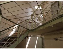 Metalltreppe und Laufgang in einem Innenraum mit industriellem Design, umgeben von hellgrünen Geländern und Gitterelementen. Oberlichter lassen Tageslicht auf die beige gestrichenen Wände fallen.