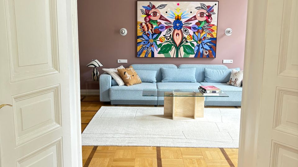 Blick durch eine weiße Flügeltür in ein helles Wohnzimmer mit Parkettboden, großem hellem Teppich, grauem Sofa und einem Glas-Couchtisch. An der roséfarbenen Wand hängt ein großes, farbenfrohes Kunstwerk; darüber eine Deckenstuckleiste und eine runde Pendelleuchte.