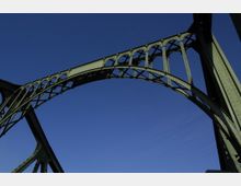 Grüne Stahlbrückenkonstruktion mit einem großen, gebogenen Bogenelement und Verstrebungen, aufgenommen gegen einen klaren blauen Himmel. Die filigranen, gerippten Strukturelemente sind deutlich erkennbar.