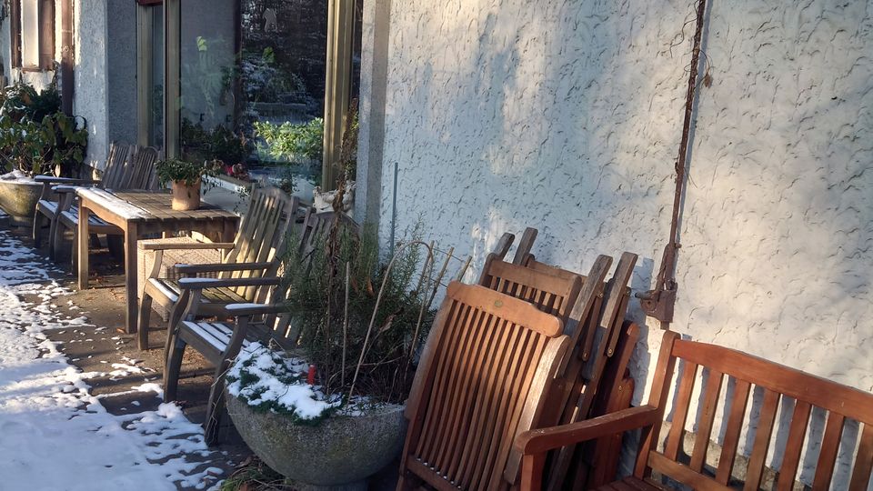 Außenbereich eines Gebäudes mit weißer Putzfassade, großen Fenstern und geschlossenen Rollläden. Vor dem Gebäude stehen Holzbänke, Tische und Stühle, teils in Schnee bedeckt; daneben eine große Pflanzschale.