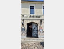 Eingangsfassade eines hellen, zweigeschossigen Gebäudes mit dem Schriftzug „Alte Schmiede“ über einem großen Rundbogenportal. Das Portal hat ein geöffnetes, schmiedeeisernes Gittertor mit Ornamenten und führt in einen gepflasterten Durchgang; seitlich hängen Schilder und ein Aushangkasten.