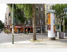 Straßenszene in einer Stadt mit mehrstöckigen Wohnhäusern, einem Baum im Vordergrund und einem runden Werbesäulenständer mit der Aufschrift „Szene Info“. Im Hintergrund ein Café mit Außenbestuhlung und weitere Geschäfte, darunter ein gelbes Gebäude mit der Aufschrift „Café Kaktus“.