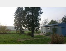 Blick auf einen ländlichen Garten mit Rasenfläche, großen Nadelbäumen und einer kleinen gepflasterten Terrasse. Rechts steht ein eingeschossiges, blau-graues Holzhaus mit Schiebetür und kleinem Fenster; dahinter sind Felder und ein Zaun zu sehen.