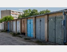 Reihe von Garagen mit unterschiedlichen farbigen Holztoren, umgeben von einer Betonfassade, im Hintergrund ein Wohngebäude und grüne Bäume. Der Boden davor ist mit Grasbüscheln und Kies bedeckt.