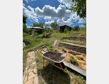 Verwilderter Garten mit Hochbeeten aus Ziegeln und einem Schubkarren voller Erde im Vordergrund, daneben ein teils überwucherter Ziegelweg. Im Hintergrund stehen ein Sonnenschirm, Bäume und eine kleine Gartenhütte unter einem Himmel mit großen Wolken.