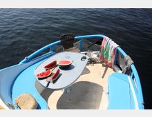 Vorderdeck eines kleinen Bootes mit blauen Relingdetails, einem Tisch mit geschnittenen Wassermelonen und einem Messer darauf. Im Hintergrund ein Stuhl, ein Handtuch über der Reling und ein Blick auf ruhiges Wasser.