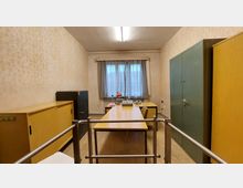 Ein einfacher Büroraum mit Holzmöbeln, darunter ein Schreibtisch mit Retro-Büroartikeln wie Schreibmaschine und Lampe. Die Wände sind mit dezent gemusterten Tapeten verkleidet, ein Fenster mit lichtdurchlässigen Vorhängen lässt Tageslicht herein, und zwei große Schränke säumen den Raum.