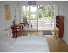 Ein helles Schlafzimmer mit einem Doppelbett im Vordergrund, einem kleinen Holztisch mit Stühlen links und einem Bücherregal rechts. Große Glastüren führen auf einen Balkon mit Blick auf grüne Bäume und einen See im Hintergrund.