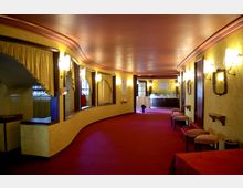 Ein geschwungener Korridor in einem Theater oder Veranstaltungsraum mit gelben Wänden, roten Teppichen und goldenen Vorhängen über Fenstern. An den Wänden hängen Spiegel und Beleuchtungskörper, im Hintergrund ist eine Barbereich mit weißem Tisch und Gläsern sichtbar.