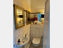 Kleines Badezimmer mit weißen Fliesen und schwarzem Akzentstreifen. Zu sehen sind ein wandhängendes WC, ein kleines Waschbecken mit Seifenspender, goldgerahmte Spiegel und dekorative Elemente wie Blumen in einer Vase und gestapelte Handtücher.