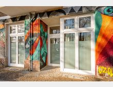 Eingangsbereich eines Gebäudes mit großen weißen Fenster- und Türelementen sowie teils blickdicht folierten Scheiben. Die Fassade ist mit farbenfrohem Graffiti und geometrischen Mustern gestaltet; davor liegt ein gepflasterter Gehweg mit Fallrohren an der Wand.