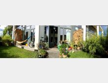 Moderne Terrasse mit Glasfront und Blick auf einen kleinen Garten. Im Vordergrund stehen zahlreiche Blumen in Töpfen, eine Hängematte, ein Fahrrad und gestapeltes Kaminholz; die Umgebung ist von grünem Rasen und Pflanzen umgeben.