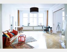 Helles Wohnzimmer mit großen Fenstern, weißen Wänden und Parkettboden. Der Raum ist mit einem braunen Ledersofa, farbigen Kissen, Teppichen, einem Schreibtisch, einer Stehlampe und einer schwarzen Hängelampe ausgestattet; ein Hund steht in der Nähe.