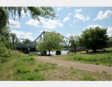 Eine grüne Parklandschaft mit einem breiten Kiesweg im Vordergrund, dahinter die Stahlkonstruktion der Glienicker Brücke, die über einen Fluss führt. Die Szene ist von Bäumen und Wiese gesäumt, mit leicht bewölktem Himmel im Hintergrund.