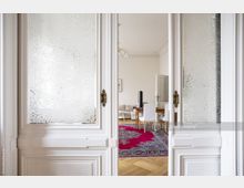 Weiße, verzierte Flügeltür mit strukturierten Glaseinsätzen, die in einen hellen Wohnraum mit Holzboden, einem großen bunten Teppich, Esstisch und Stühlen führt. Im Hintergrund sind ein Sofa und eine Zimmertür zu sehen.