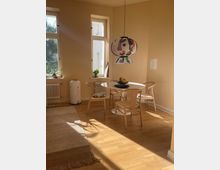 Essbereich in einem hellen Zimmer mit beigen Wänden und großen Fenstern, die Tageslicht einlassen. Ein runder Holztisch mit vier Stühlen steht auf einem Holzboden, daneben hängen eine farbige Pendelleuchte und ein Teppich im Vordergrund.