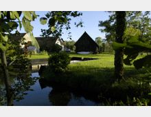 Eine ländliche Szene mit einer Wiese, einem Teich und mehreren Gebäuden, darunter ein dunkles Holzhaus und ein Gebäude mit einem Gewächshaus. Im Vordergrund fließt ein kleiner Wasserkanal, umgeben von Bäumen und Sträuchern.