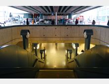 Unterirdischer Zugang zu einer U-Bahn-Station, mit einer Treppe und einem Rolltreppenbereich, flankiert von runden Lampen auf Metallpfosten. Im Hintergrund ist eine städtische Umgebung mit Verkehr, Passanten und Geschäften zu sehen.