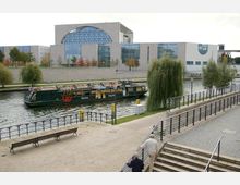 Eine moderne Architekturkulisse mit dem Berliner Regierungsviertel im Hintergrund, einer breiten Wasserstraße im Vordergrund, auf der ein Touristenboot fährt. Am Ufer befinden sich Weidenbäume und ein gepflasterter Gehweg mit Sitzbänken sowie Geländer.