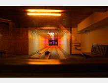 Kegelbahn in einem geschlossenen Raum mit Holzverkleidung an den Wänden, einer beleuchteten Decke und zwei Kegelbahnen, die zu einem lebhaft beleuchteten Kegelstand führen. Im Vordergrund sind Sitzbänke und ein Waschbecken an der Wand sichtbar.