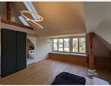 Modernes Dachgeschosszimmer mit schrägen Decken, sichtbaren Holzbalken und hellem Holzboden, beleuchtet durch große Fensterfront und ein Dachfenster. Unter den Fenstern verläuft eine lange Holzkommode; links steht ein hoher, dunkler Schrank und daneben führt eine Tür in ein Badezimmer.