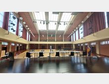 Großer Veranstaltungssaal mit Holzfußboden, hohen Decken und einer Glasdecke, die viel Tageslicht hereinlässt. Im Vordergrund eine Bühne mit Stühlen und Tischen, im Hintergrund Reihen von Tischen und Stühlen sowie rote Vorhänge entlang der Fensterwände.