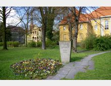 Grüne Parklandschaft mit einem Denkmal aus Stein, das die Inschrift „Den Opfern 1933-1945“ trägt, umgeben von Bäumen und Blumenbeeten. Im Hintergrund sind gelbe Gebäude mit roten Dächern sowie eine Kirche mit spitzem Turm zu sehen.
