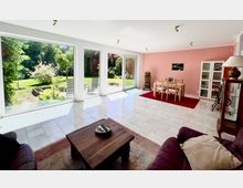 Heller Wohn- und Essbereich mit großen bodentiefen Fenstern und Glasschiebetür zur Terrasse und in einen grünen Garten. Fliesenboden, rosa Akzentwand sowie Esstisch auf rotem Teppich und Wohnmöbel im Vordergrund.