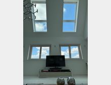 Modernes Wohnzimmer mit weißen Wänden und hohen Decken, durchbrochen von vier großen Dachfenstern, die einen klaren Blick auf den blauen Himmel mit Wolken ermöglichen. Im unteren Bereich befindet sich ein Fernseher auf einem schlichten weißen TV-Schrank vor zwei rechteckigen Fenstern.