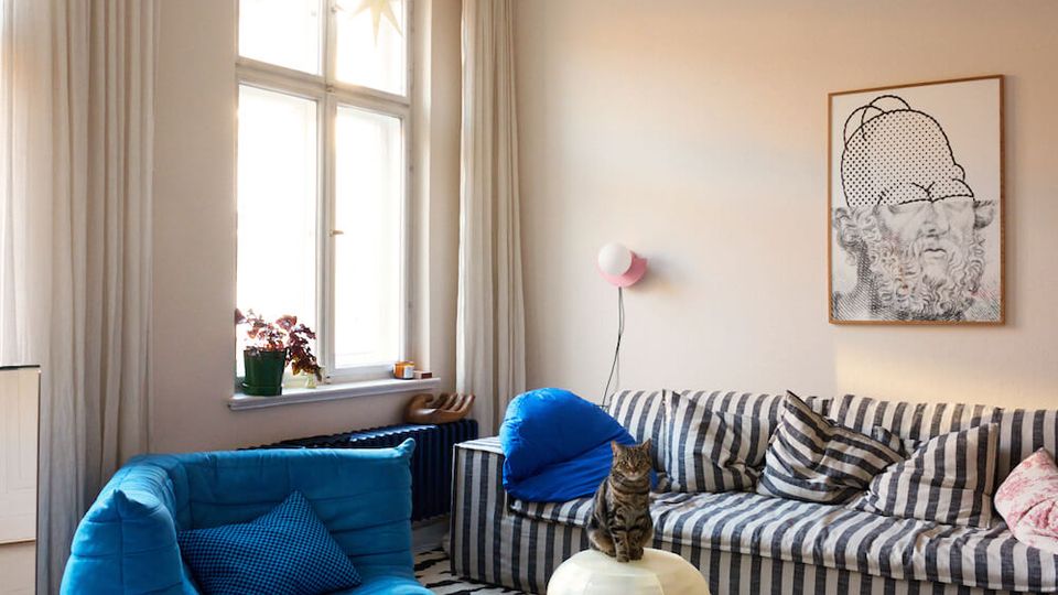 Bright living room with high ceilings, ornate crown molding, and a tall window framed by long curtains. The space features a striped sofa, a blue lounge chair, a zebra-pattern rug, and a small round translucent coffee table beneath a framed wall artwork.