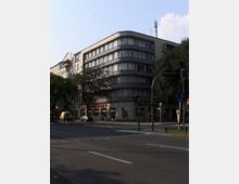 Eckgebäude mit abgerundeter Fassade und großen Fenstern im Stil der Nachkriegsmoderne, an einer Kreuzung mit Bäumen, Verkehrsampeln und Gehwegen. Im Erdgeschoss befinden sich Geschäfte mit Schaufenstern.