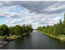 Ein gerader Kanal mit ruhigem Wasser, gesäumt von dicht bewachsenen grünen Bäumen auf beiden Ufern, unter einem teils bewölkten Himmel.