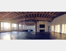 Großer, lichtdurchfluteter Raum mit Betondecke und großen Fensterfronten, die viel Tageslicht hereinlassen. Der Raum ist minimal eingerichtet, mit wenigen Möbelstücken wie Sofas und einer kleinen Küchenzeile an einer der Wände.