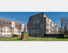 Mehrfamilienhäuser in einem Wohngebiet mit einfacher Architektur, darunter ein Gebäude mit grauer Fassade und Balkonen sowie eines mit einem roten Ziegeldach. Im Vordergrund steht ein bemalter Stromkasten auf einer Rasenfläche, umgeben von kahlen Bäumen und einem Metallzaun.