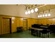 Ein historischer Raum mit Jugendstil-Elementen, gekennzeichnet durch gelblich-braune Wandvertäfelungen mit dekorativen Mustern und verzierten Holztüren. Die Decke ist mit hellen Kugelleuchten und dekorativen Metallstreben gestaltet, während der Raum mit einfachen schwarzen Möbeln und Stühlen ausgestattet ist.