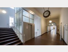 Treppenhaus und Flur in einem historischen Gebäude mit braunem Bodenbelag, einem Aufzug mit Milchglasverkleidung sowie einer großen, hängenden Uhr. Der Flur ist durch Reihen von Rundbogenfenstern hell erleuchtet und mit Kugellampen ausgestattet.