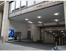 Einfahrt zu einem öffentlichen Parkhaus mit der Beschriftung „Q-Park Opern Carree“. Die Einfahrt verfügt über moderne Beleuchtung, eine Schranke, ein Hinweisschild mit Parkgebühren und Richtungsangabe sowie eine Rampe, die in den Parkplatz führt.