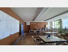 Großer Pausen- oder Aufenthaltsraum mit Holzvertäfelung, Parkettboden und abgehängter Rasterdecke, ausgestattet mit mehreren Tischen und Stühlen. Links hängen große Whiteboards und Stellwände, rechts sorgen große Fenster mit Pflanzen am Rand und eine kleine Küchen- bzw. Getränkezeile im Hintergrund für Tageslicht und Versorgung.
