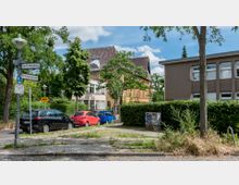 Straßenecke mit einem Straßenschild „Am Fichtenberg / Rothenburgstraße“, geparkten Autos und umliegender Vegetation. Im Hintergrund ein gelbes Gebäude mit historischer Architektur, rechteckige Fenster sowie ein modernes, graues Gebäude mit flachen Fenstern.
