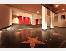 Eingangsbereich eines Veranstaltungsorts mit rotem Teppich auf einer erhöhten Bühne und roten Vorhängen im Hintergrund. Auf dem schwarzen Boden sind Sterne mit Namen eingebettet, ähnlich dem Walk of Fame.